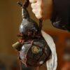 A person holding an artisanal ceramic gourd water bottle with a dark, kiln-fired glaze, secured in a hand-tied brown rope carrier with a small brass bell.