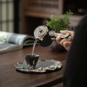 A person mindfully pouring tea from an artisanal side-handle handmade ceramic teapots into a matching wabi-sabi style cup on a lotus leaf tray.