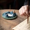 A person meditating in front of the mountain style backflow incense burner, which is creating a calming smoke waterfall on a wooden table.