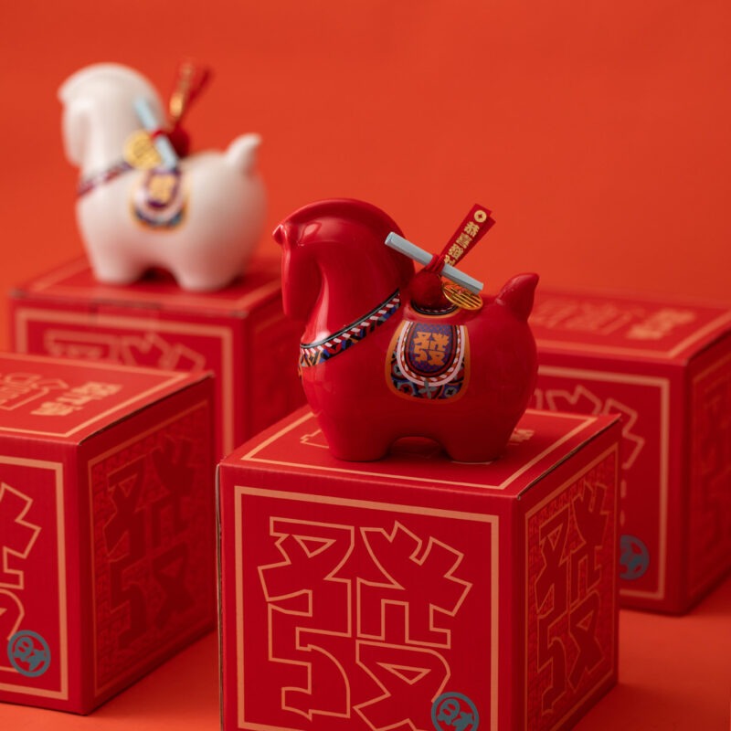 Close up of a red year of the horse ceramic ornament showing the golden coin detail, greeting scroll, and glossy porcelain finish for Lunar New Year decoration.